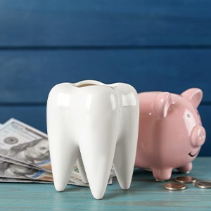 A ceramic tooth model next to a piggy bank, dollar bills, and coins